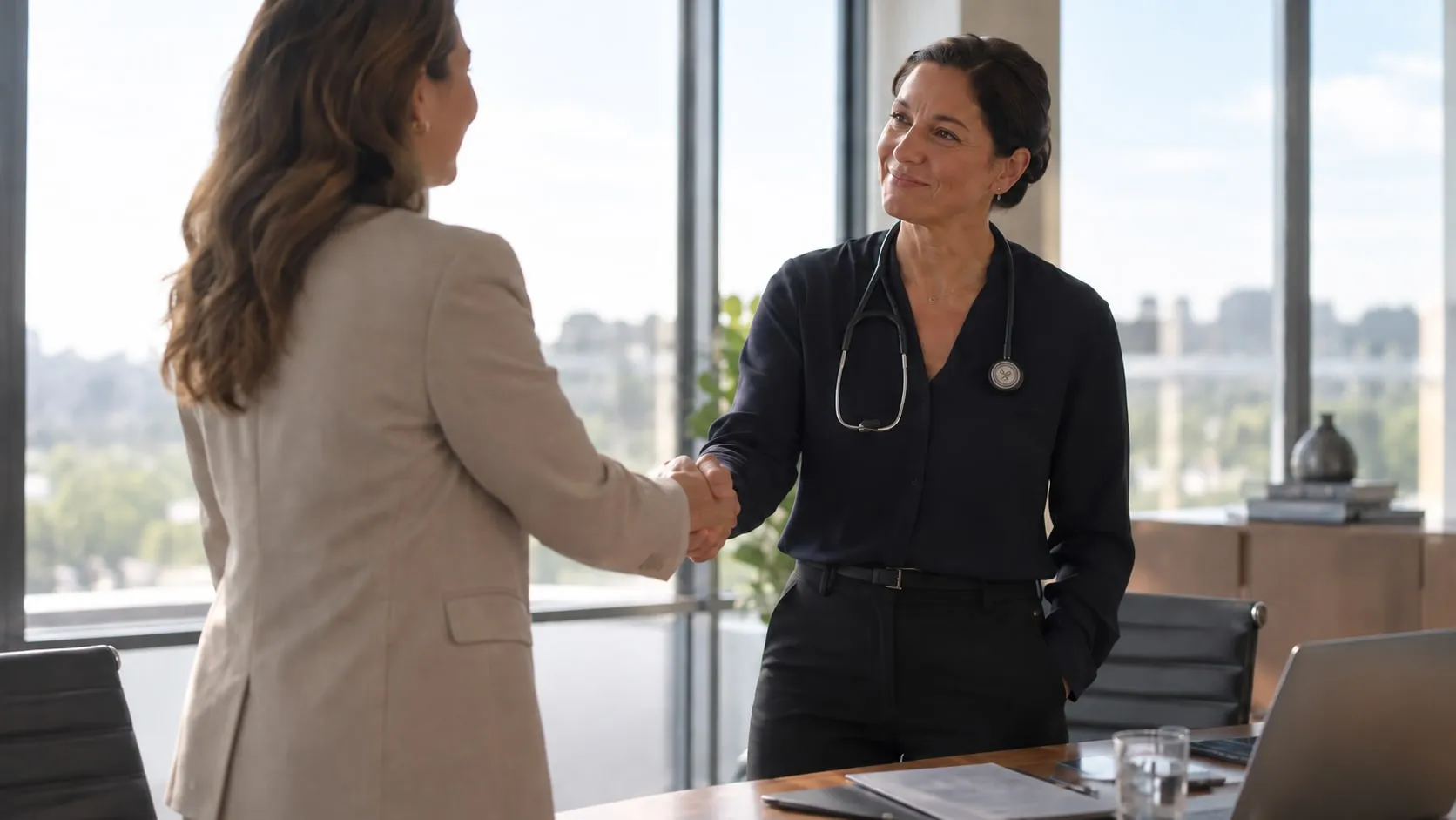 A clinician wearing a stethoscope shakes hands with a client across a boardroom table